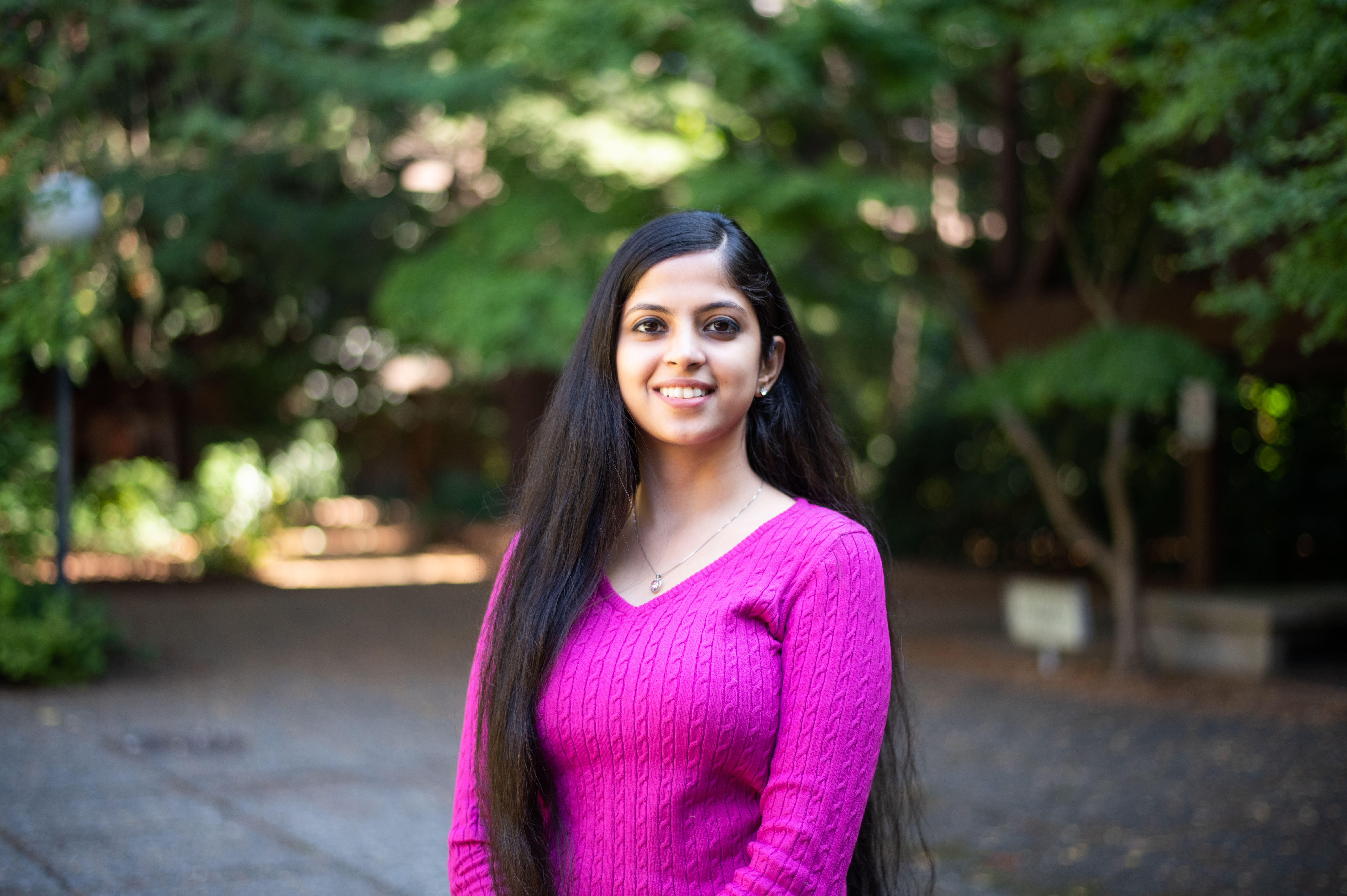 A photo of Anukriti, an asian woman who is smiling at the camera. She is wearing a pink sweater and a pendant around her neck. She has long, black hair parted to one side. The background is blurry, with green trees, indicating this photo was taken outdoors.